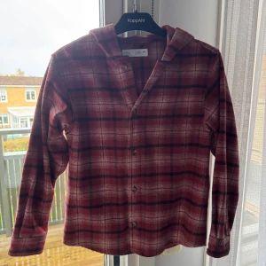 Rutig röd skjorta med huva från Zara - Rutig skjorta från Zara i rött och svart med huva och knappar framtill. Skjortan har lång ärm och är tillverkad i mjukt material som känns skönt mot huden. Perfekt för din son som gillar avslappnad stil med lite edge.