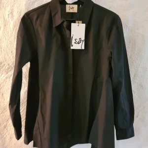 Svart skjorta från Isay, strl 34 - Stilren svart skjorta från Isay i 100% bomull. Klassisk krage, långa ärmar och dolda knappar framtill. Modellen heter Bellis New Shirt och är utsvängd nedtill och lite längre ibak, funkar till många olika looks. Perfekt för dig som gillar enkla och snygga basplagg.