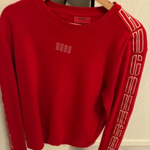 Röd sweatshirt från Hugo med tryck - Riktigt snygg röd sweatshirt från Hugo med coolt vitt tryck på bröstet och längs båda ärmarna. Tröjan har rund hals och lång ärm, perfekt för dig som gillar streetstyle och vill sticka ut lite extra. Materialet är mjukt och skönt, troligen bomull.
