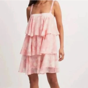 Rosa volangklänning med knyt - Supersöt kortklänning i ljusrosa och vit med volanger i flera lager och smala axelband. Klänningen har ett mjukt, flowigt tyg och ett knytband framtill. Perfekt för sommarens alla tillfällen när du vill känna dig extra fin.