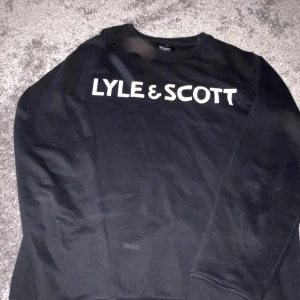 Svart sweatshirt från Lyle & Scott - Svart sweatshirt från Lyle & Scott med stor vit logotyp på bröstet. Tröjan har rund hals, långa ärmar och ribbade muddar vid ärmslut och nederkant. Perfekt för dig som gillar enkel och stilren streetwear.