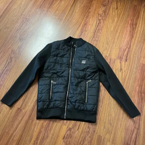 Svart quiltad bomberjacka från BOSS - Svart bomberjacka från BOSS med quiltad framsida och släta ärmar och rygg. Jackan har två fickor med dragkedja, ribbad krage och nederkant. Materialmix med polyester och stickad textil ger en clean och modern look. Perfekt för dig som gillar stilrena detaljer.