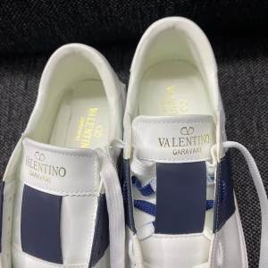 Valentino Garavani vita sneakers - Snygga vita sneakers från Valentino Garavani med breda mörkblå detaljer på sidorna och vita snören. Skorna har en rund tå, platt sula med mönstrad undersida och är tillverkade i skinn. Perfekt för dig som gillar stilrena och lyxiga sneakers med exklusiv känsla.