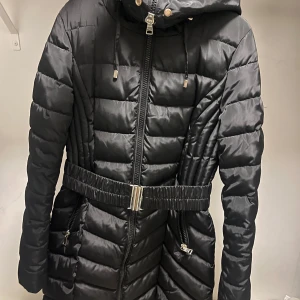 Svart pufferjacka med leopardfoder - Svart pufferjacka från Sustans med snyggt leopardmönstrat foder och orange kant. Jackan har huva, dragkedja framtill, två fickor med dragkedja och ett brett bälte i midjan med metallspänne. Materialet är syntet och jackan är quiltad för extra värme. Pris går att diskutera vid snabba affärer