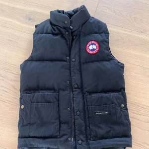 Svart dunväst från Canada Goose - Svart dunväst från Canada Goose storlek xs med klassisk logga på bröstet. Västen har hög krage, två stora fickor med tryckknappar och dragkedja framtill. Yttermaterialet är polyester och bomull, fodrad med dun för extra värme. Perfekt för kyliga dagar och snygg att styla över hoodie. Mycket bra skick men lite solblekt.