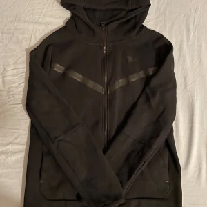 Svart Nike tech med huva  - Svart zip från Nike i storlek S med hel dragkedja, huva och diskreta svarta detaljer över bröstet. Tröjan har två fickor framtill och en extra ficka på ärmen. Perfekt för dig som gillar sportig och clean stil.