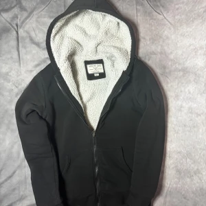 Svart hoodie från Denim Day med teddyfoder - Svart hoodie från Denim Day med dragkedja och mysigt vitt teddyfoder i hela insidan och huvan. Klassisk modell med huva och två fickor framtill. Perfekt för kyliga dagar när du vill vara både varm och stilren.