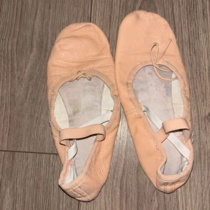 balettskor i skinn - Säljer ett par klassiska balettskor i beige skinn med rund tå och platt sula. Skorna har elastiska band över foten för extra stöd och en mjuk innersula. Perfekta för dans eller balettträning, med flexibel passform och stilren look. Det är en vatten fläck på en av skorna. De är köpta för 295.