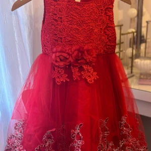 Röd spetsklänning med blommor, stl 130 - Superfin röd klänning med spetsliv och voluminös tyllkjol. Dekorerad med stora tygblommor och broderade detaljer i midjan samt längs kjolkanten. Klänningen har dragkedja bak och är ärmlös, perfekt för speciella tillfällen.