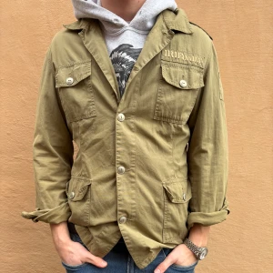 005 khakigrön overshirt  - En khakigrön parkajacka från Irie Daily med coola detaljer såsom axelklaffar och fyra stora fickor med knappar. Jackan har en klassisk krage och tryck på bröstet. Perfekt overshirt för dig som gillar streetwear och vill ha en snygg jacka nu för hösten.