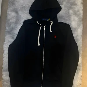 Svart hoodie från Polo Ralph Lauren XL - Svart hoodie från Polo Ralph Lauren i storlek XL. Klassisk design med vit snörning i huvan, dragkedja framtill och röd broderad logga på bröstet. Perfekt för chill dagar och streetstyle. Mjuk bomullskänsla och långärmad modell.