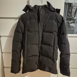 Svart pufferjacka från Canada Goose - Säljer en svart pufferjacka från Canada Goose med huva och stora fickor framtill. Jackan har quiltad design, dragkedja och tryckknappar. Perfekt för kalla dagar och har en klassisk look med diskret logga på ärmen.
