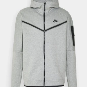 Nike tech - ljusgrå Nike tech med svart logga på bröstet och hel dragkedja framtill. Tröjan har huva, långa ärmar och svarta detaljer längs dragkedjor och fickor. Nypris 1300kr