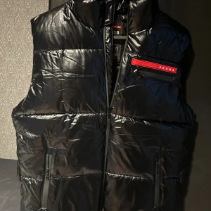 Svart dunväst från Prada - Snygg svart dunväst från Prada med glansig finish och hög krage. Västen har flera dragkedjor, inklusive en bröstficka med röd Prada-logga. Perfekt för lager-på-lager och streetstyle. Stilsäker och modern look med quiltad design.