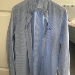 Blårandig skjorta från Lacoste - Snygg blårandig skjorta från Lacoste i slim fit-modell. Skjortan har klassisk krage, långa ärmar och knäppning framtill. Tillverkad i bomull och prydd med den ikoniska krokodilloggan på bröstet. Perfekt för en clean och fräsch look.