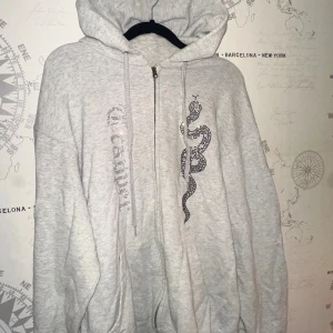 Grå hoodie med strass och orm - Säljer en ljusgrå hoodie från Trapper med dragkedja, huva och fickor. På framsidan finns en stor orm i svarta strasstenar och texten 'Trapper' i glittriga stenar. Oversized passform, storlek Large