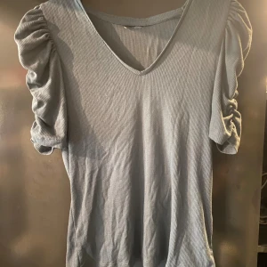 Ljusblå ribbad topp med puffärmar - Snygg ljusblå ribbad topp med v-ringning och korta puffärmar. Toppen har en figurnära passform och är perfekt för dig som gillar trendiga detaljer. Materialet känns mjukt och stretchigt.