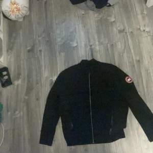 Svart jacka från Canada Goose - Säljer en svart jacka från Canada Goose med klassisk logga på ärmen. Jackan har hel dragkedja framtill, ståkrage och två fickor med dragkedja. Perfekt för dig som vill ha en stilren och ikonisk look.