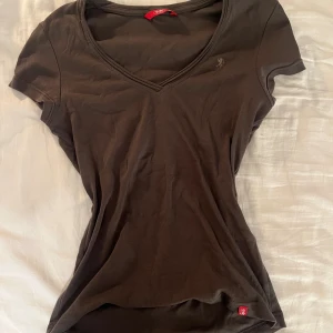 Brun v-ringad topp från EDC by Esprit - Säljer en brun, figurnära topp från EDC by Esprit i mjuk bomull. Toppen har v-ringad hals, korta ärmar och en liten logga broderad på bröstet. Perfekt för dig som gillar enkel och stilren design med en skön passform.