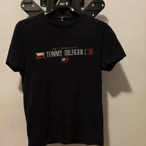 Svart Tommy Hilfiger t-shirt med broderi - Svart t-shirt från Tommy Hilfiger med broderad logga och flaggor i rött, vitt och guld på bröstet. Klassisk rund hals och korta ärmar. Materialet är mjuk bomull som känns skönt mot huden. Perfekt för dig som gillar stilrena och ikoniska plagg.