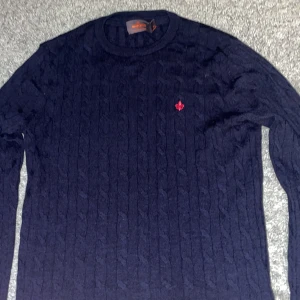 Morris Cableknit storlek XS - Snygg marinblå kabelstickad tröja från Morris i 100% merinoull. Tröjan har rund hals och ett rött broderat emblem på bröstet. Perfekt för dig som gillar klassisk stil med en modern twist. Passar dig som vill ha en mjuk och varm tröja med cool detalj.