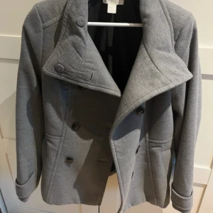 Kappa med hög krage  - Stilren grå kappa från H&M med hög krage och dubbelknäppning framtill. Jackan har två sidfickor och långa ärmar