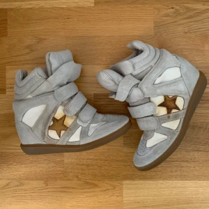 Isabel Marant wedge sneakers grå 37 - Säljer ett par ikoniska Isabel Marant wedge sneakers i grå mocka och vitt skinn med bruna stjärndetaljer på sidorna. Skorna har tre kardborreband, rund tå och gummisula. Perfekta för dig som vill ha en cool och sportig look med extra höjd. På sista bilden syns en defekt som man behöver fixa till hos skomakaren.