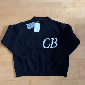 Svart stickad tröja Cole Buxton - Svart stickad tröja från Cole Buxton med vit CB-logga på bröstet. Tröjan har rund hals, ribbade muddar och långärmad passform. Enkel och stilren design som funkar till många olika looks. Perfekt för dig som gillar cleana och snygga plagg.
