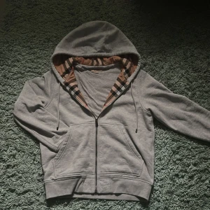 Grå hoodie från Burberry med rutig insida - Grå hoodie från Burberry med klassisk rutig insida. Tröjan har huva med snörning. Perfekt för dig som gillar  stilrena och lyxiga streetwear-plagg.  Storlek M