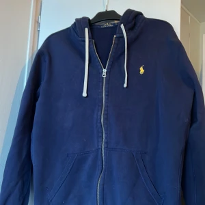 Marinblå hoodie från Polo Ralph Lauren - Säljer en marinblå hoodie från Polo Ralph Lauren med dragkedja och huva. Tröjan har vita snören, två fickor framtill och en gul broderad logga på bröstet. Perfekt för en avslappnad stil.