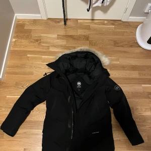 Svart dunjacka från Canada Goose M - Säljer en svart dunjacka från Canada Goose med avtagbar päls på huvan. Jackan har lång ärm, dragkedja och klassisk svart patch på ärmen. Perfekt för kalla vinterdagar och har en clean, stilren look. Fodrad med dun och har flera praktiska fickor.