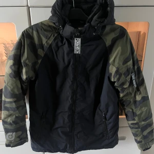 Skidjacka/vinterjacka  stl 158 WOXO 720 - Säljer en svart vinterjacka från WOXO 720 med camouflagemönstrade ärmar och huva. Jackan har dragkedja framtill, snörning vid huvan och praktiska fickor med dragkedja. 