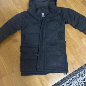 Svart dunjacka från Canada Goose - Svart dunjacka från Canada Goose med huva och stora fickor framtill. Jackan har klassisk patch på ärmen, dragkedja och knappar framtill samt quiltad design. Perfekt för kalla vinterdagar och riktigt varm. Snygg och funktionell stil, päls med. Vid snabb affär så kan jag gå ner med priset 