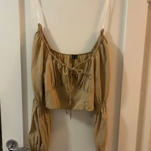 Beige croppad blus från Vero Moda - Säljer en beige croppad blus från Vero Moda med långa puffärmar och smockdetalj i midjan. Toppen har snörning framtill och en luftig, avslappnad passform. Perfekt för dig som gillar trendiga och bekväma plagg med snygga detaljer.