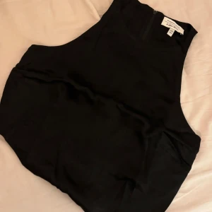 Svart ärmlös topp från & Other Stories - Säljer en svart, stilren ärmlös topp från & Other Stories. Toppen har hög halsringning och dragkedja i ryggen för en clean look. Perfekt att styla med både jeans och kjol. Materialet är mjukt och har en lätt glansig finish.