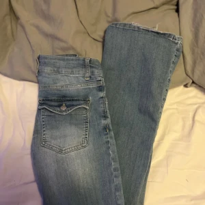 Ljusblå bootcut jeans från Nelly - Säljer ett par ljusblå bootcut jeans från Nelly med slitna detaljer vid knäna. Jeansen har klassisk femficksdesign, bälteshällor och dragkedja med knapp framtill. Perfekta för en avslappnad och trendig look.