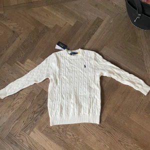 Vit stickad tröja från Polo Ralph Lauren - Vit stickad tröja från Polo Ralph Lauren med klassiskt kabelstickat mönster och broderad logga på bröstet. Rund hals och långa ärmar, perfekt för en clean och stilren look. Ribbstickade muddar vid ärmslut och nederkant. Passar någon runt 173