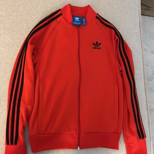 Röd Adidas track jacket med svarta ränder - Riktigt snygg röd Adidas track jacket med klassiska svarta tre ränder längs ärmarna och broderad logga på bröstet. Jackan har dragkedja hela vägen, ståkrage och två fickor. Perfekt för en sportig och retro vibe.