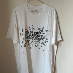 Maison Margiela vit t-shirt med print - Maison Margiela t-shirt i vit bomull med svart splatter-print och siffror på framsidan. Klassisk rund hals och korta ärmar. Snygg och unik design som sticker ut, tillverkad i Italien. Perfekt för dig som gillar statement-plagg och exklusiva märken.