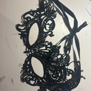 Svart spetsmask med knytband - Säljer en svart maskeradmask i spets med snygga, detaljerade mönster och knytband i satin. Masken har en elegant och mystisk look, perfekt för maskerad eller bal. Den är lätt och sitter bekvämt runt huvudet tack vare de breda banden.
