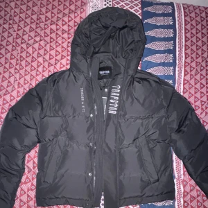 Svart pufferjacka från Trapstar XL - Svart pufferjacka från Trapstar med broderad logga på bröstet och stor Trapstar-text på ryggen. Jackan har huva, dragkedja och tryckknappar framtill. Oversized passform och cool streetstil, perfekt för kalla dagar.