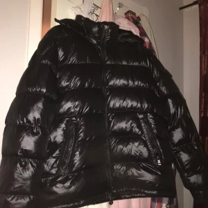 Svart Moncler pufferjacka med huva - Säljer en svart pufferjacka från Moncler med glansig finish och huva. Jackan har dragkedja framtill, två fickor med dragkedja och Moncler-logga på ärmen. Perfekt för kalla dagar och riktigt snygg streetstil.