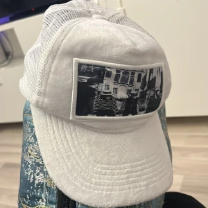 Vit emilio pendeza keps i bra skick  - Säljer en vit keps med meshpaneler och böjd skärm. Framtill sitter en svartvit patch med ett coolt foto-tryck. Kepsen har klassisk trucker-stil och är perfekt för dig som gillar streetwear och vill sticka ut med en unik detalj.