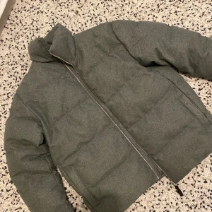 Grå pufferjacka med dragkedja - Säljer en grå pufferjacka i stilren design med hög krage och hel dragkedja framtill. Jackan är vindtät och vattenavvisande, perfekt för kyliga dagar. Materialet känns mjukt och har en diskret textur. Två sidofickor och lång ärm för extra komfort.