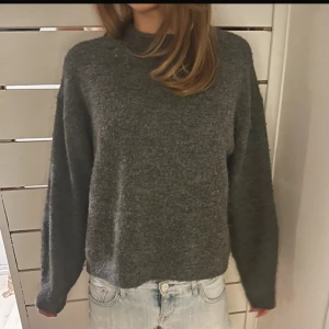 Mörkgrå stickad tröja med hög hals - Mysig mörkgrå stickad tröja med hög hals och långärmad passform. Tröjan har en loose fit och är perfekt för kyliga dagar. Enkel och stilren design som funkar till jeans eller andra favoritplagg.