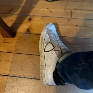 Phillip models  - Säljer ett par vita sneakers med svart häl och svart sula. Skorna har snörning och en unik detalj på sidan. Klassisk rund tå och platt sula, tillverkade i skinnliknande material. Perfekta för dig som gillar stilrena och enkla sneakers.