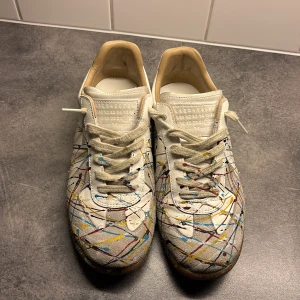 Maison Margiela sneakers med färgstänk - Unika vita sneakers från Maison Margiela med coola färgstänk i blått, gult, rött och svart. Skorna har snörning, rund tå och är tillverkade i skinn och mocka. Perfekta för dig som vill sticka ut med en konstnärlig vibe.