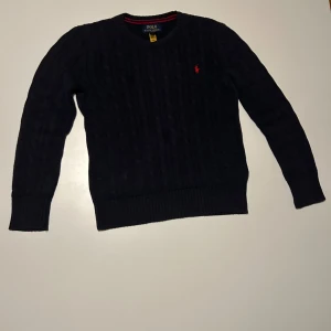 Svart kabelstickad tröja Polo Ralph Lauren - Kabelstickad svart tröja från Polo Ralph Lauren med rund halsringning och röd broderad logga på bröstet. Klassisk modell med ribbade muddar och nederkant. Perfekt för dig som gillar stilrena och tidlösa plagg.