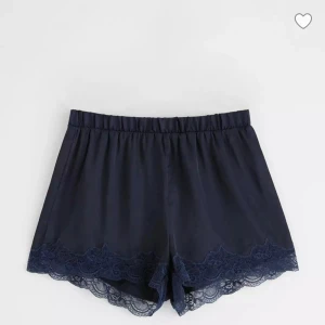 Mörkblå shorts med spetskant i satin  - Snygga mörkblå shorts med elastisk midja och dekorativ spetskant längst ner. Perfekta för dig som gillar en mjuk och feminin stil. Materialet känns lätt och skönt mot huden, och spetsen ger en extra touch av elegans.