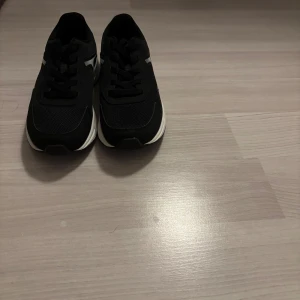 Gym / löpning skor  - Säljer ett par svarta sneakers med vit sula och snygga vita detaljer på sidan. Skorna har snörning och rund tå, tillverkade i syntetmaterial med mesh för extra komfort. Perfekta för dig som gillar stilrena och sportiga skor.